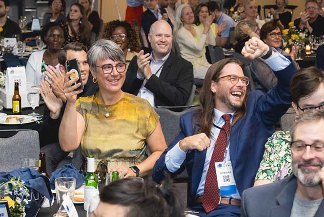 People seated at a formal event clapping and cheering with drinks and flowers on tables.