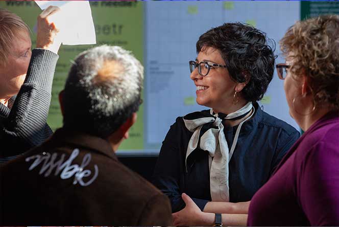 Four people sitting together engaged in discussion, with a woman in glasses smiling and looking to the side, a person holding up a paper, and a screen with charts in the background.