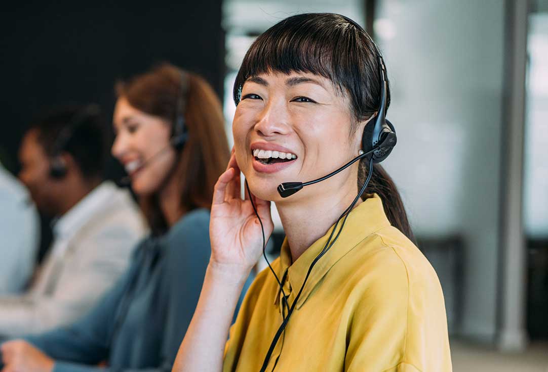 Smiling woman wearing a yellow shirt and headset, working in a newsroom support office with colleagues in the background.