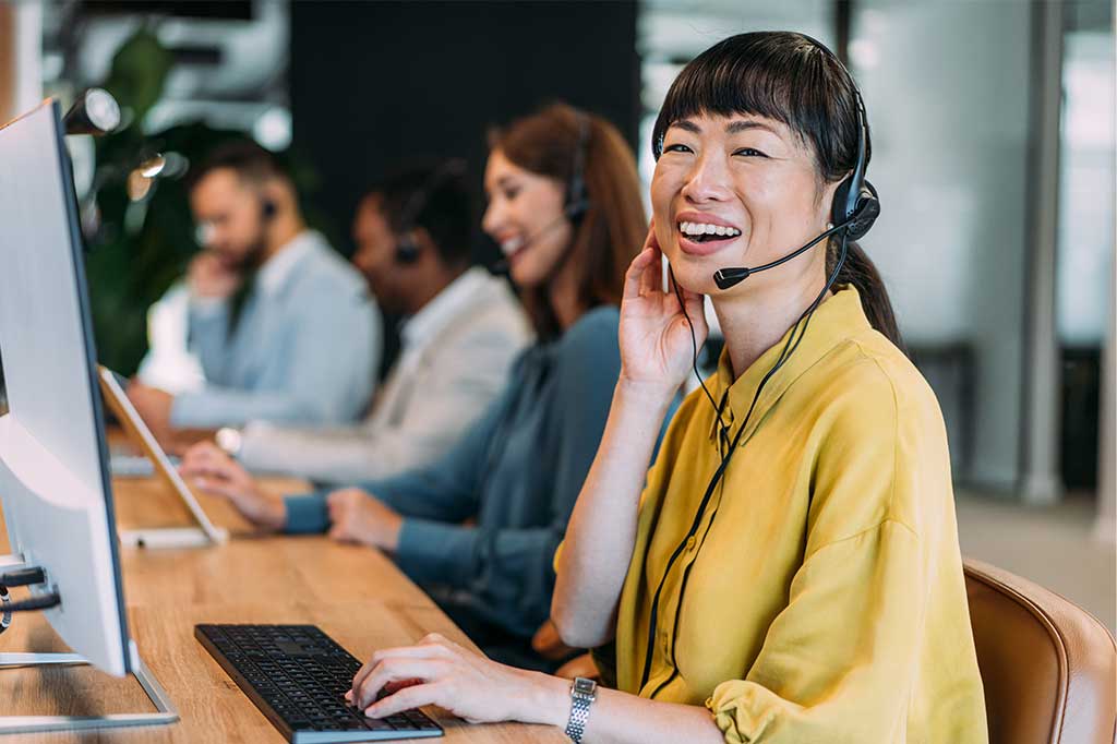 Smiling female customer support representative wearing a headset and working at a computer with colleagues in the background.
