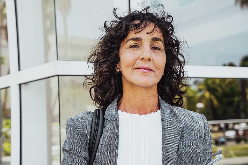 Confident woman with curly dark hair wearing a gray blazer and white blouse standing outdoors near glass windows.