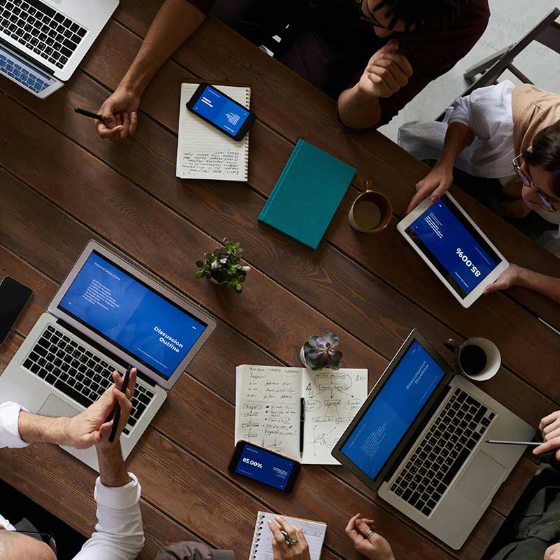 Overhead view of diverse people collaborating at a wooden table with laptops, tablets, smartphones, notebooks, and coffee cups.