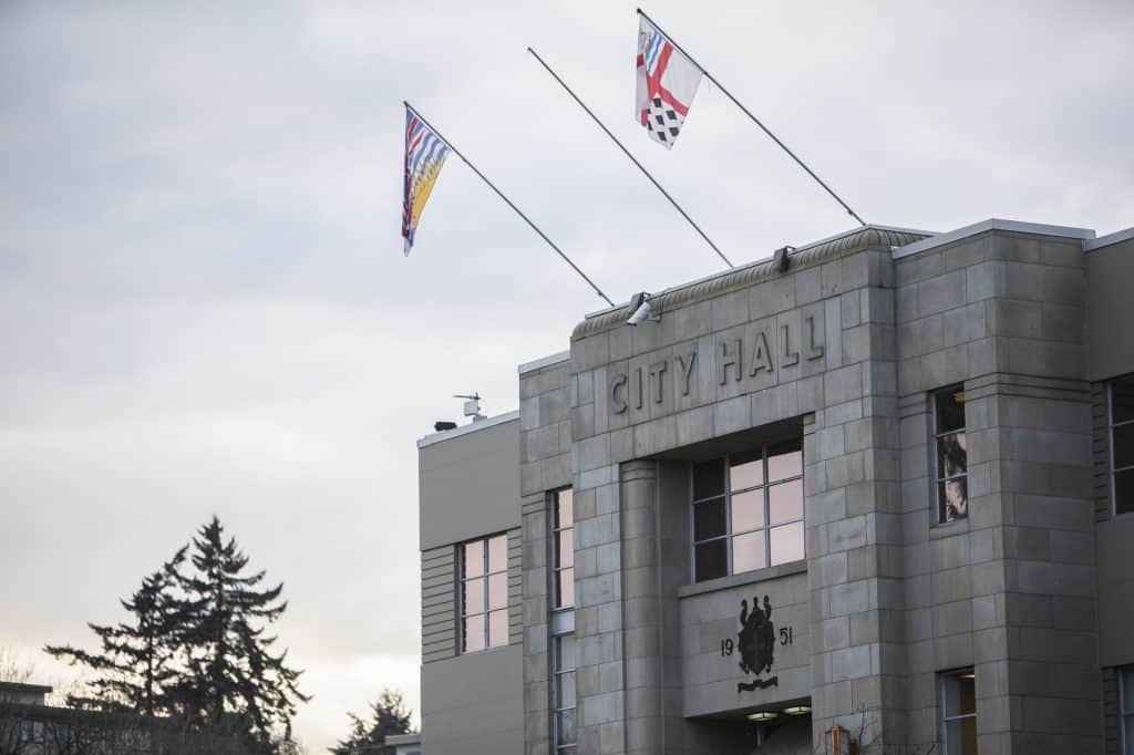 A stone building labeled "City Hall" in the City of Nanaimo displays two flags on poles and an emblem on its facade.