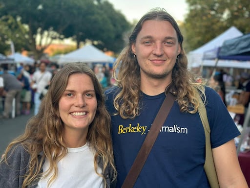Two people with long, brown hair at a street festival smiling at the camera.