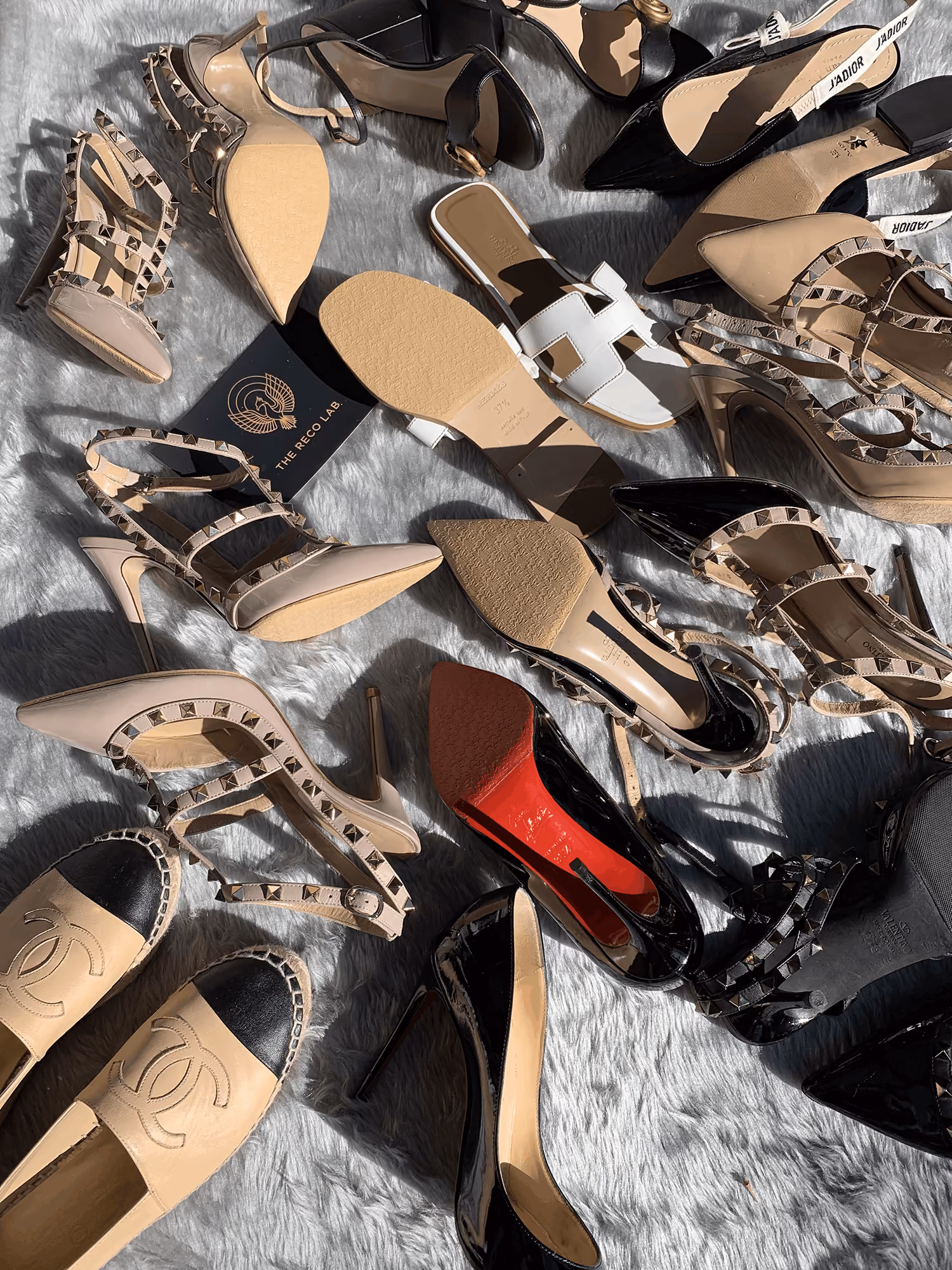 Collection of designer shoes ready for restoration, including Valentino Rockstud heels, Chanel espadrilles, Hermès Oran sandals, Dior slingbacks and Christian Louboutin red-sole heels, arranged before treatment by The Reco Lab.