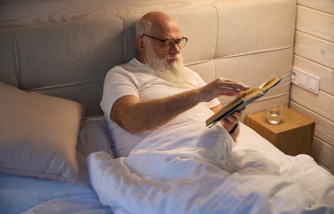 An elderly white man with short white hair and a long white beard wearing glasses and a white tee shirt reads in bed.