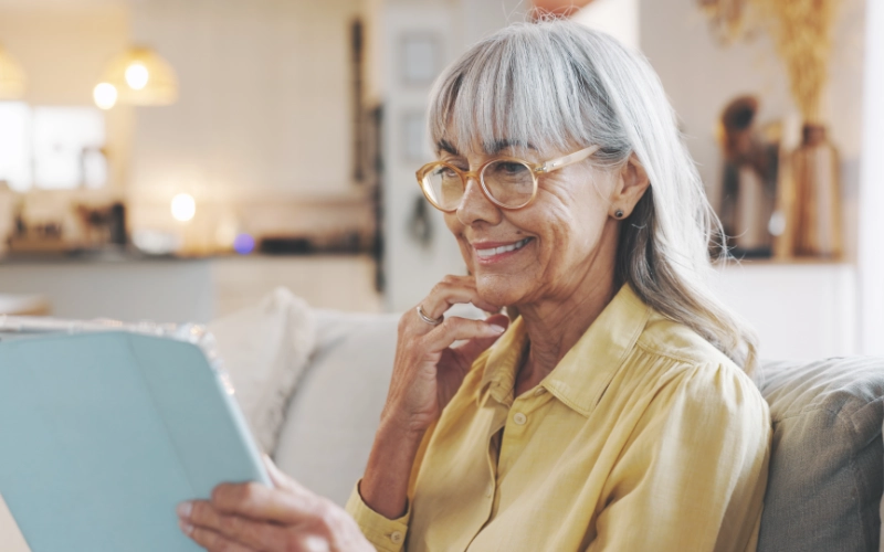 An elderly white woman with long silver hair wearing a gold blouse and looks at her tablet device and smiles warmly.