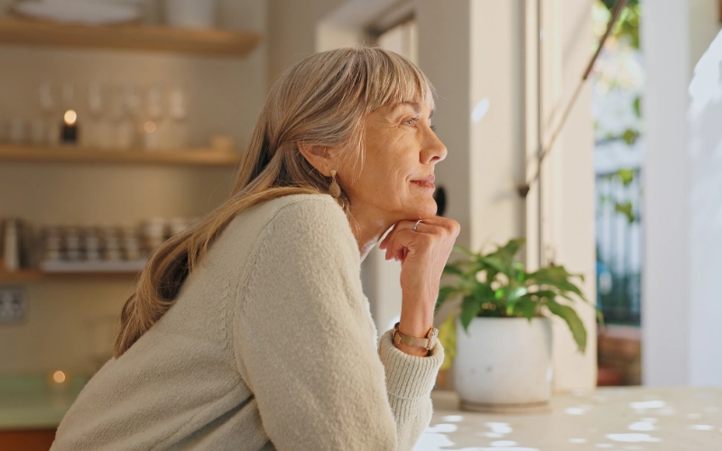 An elderly white woman with long silver hair wearing a beige sweater rests her chin on her hand and looks out the window with a pensive expression.