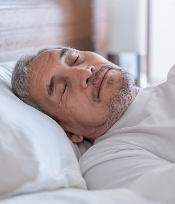 An elderly Asian man with short hair and a goatee sleeps peacefully in a white shirt and white sheets.