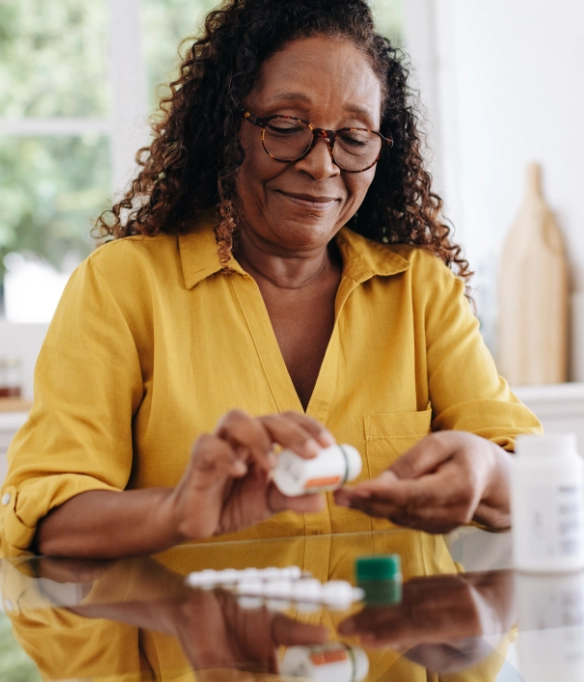 An elderly black woman with long curly hair wearing a yellow shirt and glasses takes her medications.