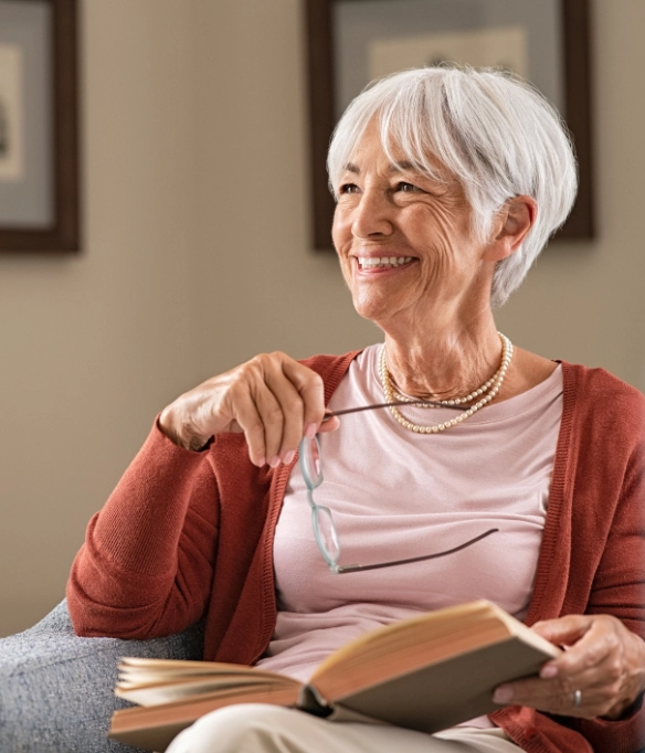 An elderly white woman with white hair in a pixie cut looks up and smiles off camera while reading a book.