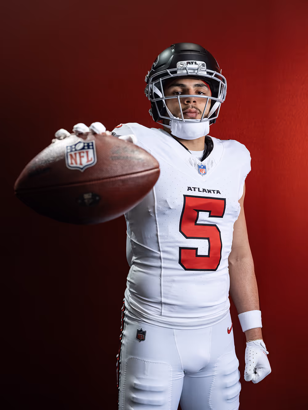 Football player wearing a white Atlanta jersey with number 5 and black helmet holding a football towards the camera against a red background.