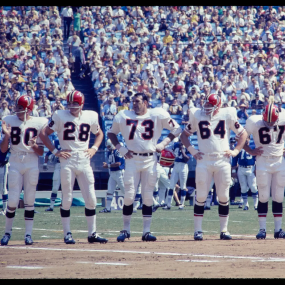 Five football players in white uniforms and red helmets standing side-by-side on a field in front of a large crowd.
