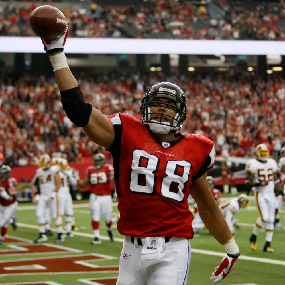 Football player in red and white Atlanta Falcons uniform raising a football in celebration on the field.