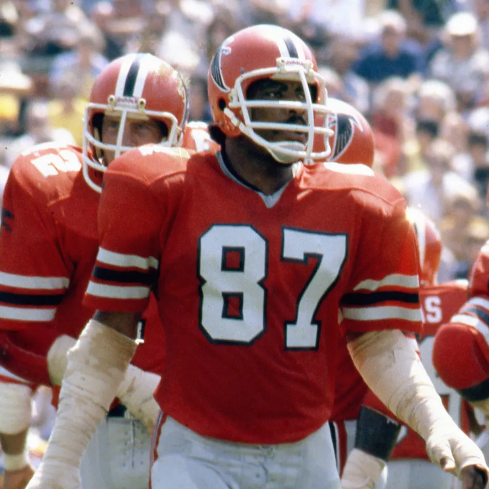 American football players wearing red jerseys with white numbers and orange helmets with a black falcon logo during a game.