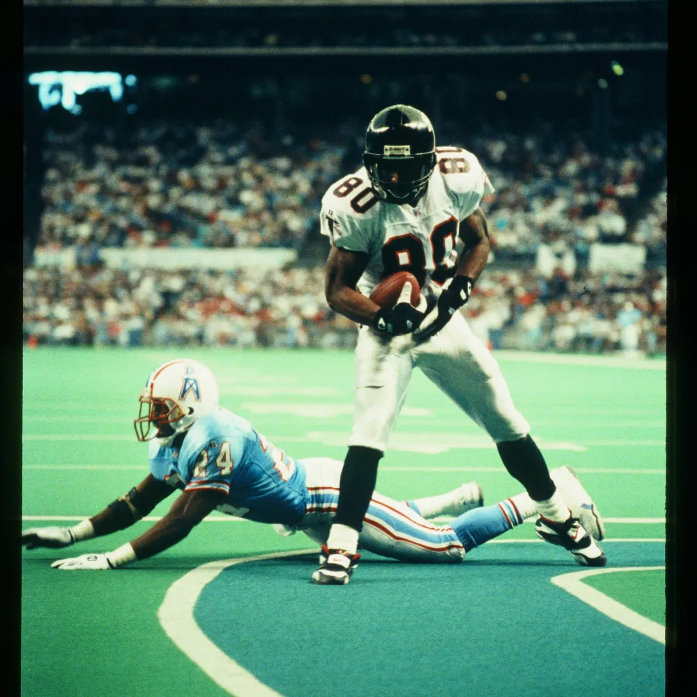 Falcons football player in white uniform with number 80 holding the ball, standing over a Tennessee Titans player lying face down on the field.