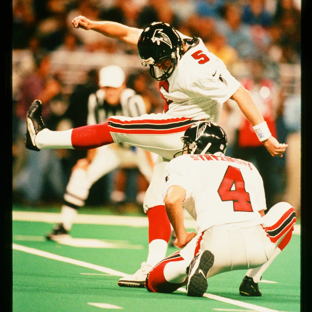 American football player in white Atlanta Falcons uniform kicking a football held by a teammate on a green field.