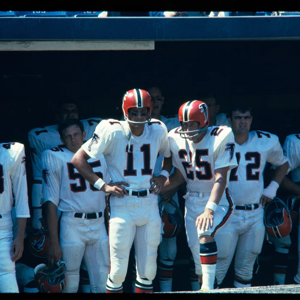 Atlanta Falcons football players in vintage white uniforms and red helmets standing in a shaded dugout.