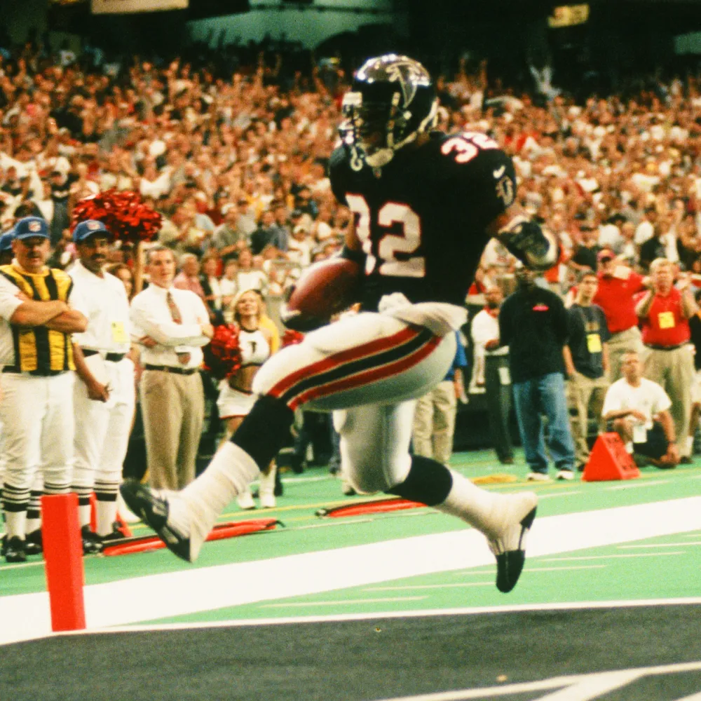 Atlanta Falcons player wearing number 32 jumping to score a touchdown in a packed football stadium.