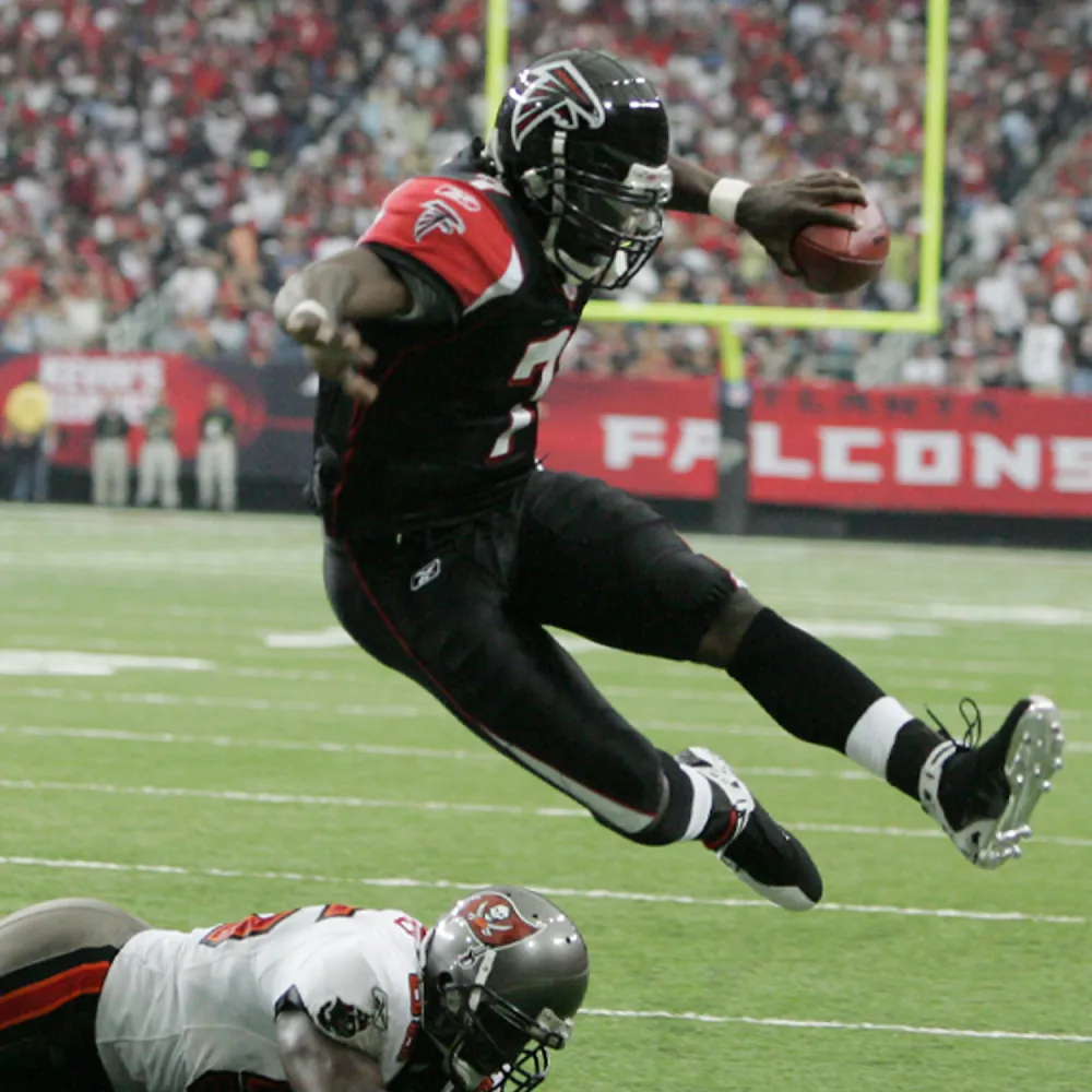 Atlanta Falcons player in black uniform leaping over a Tampa Bay Buccaneers defender on a football field.