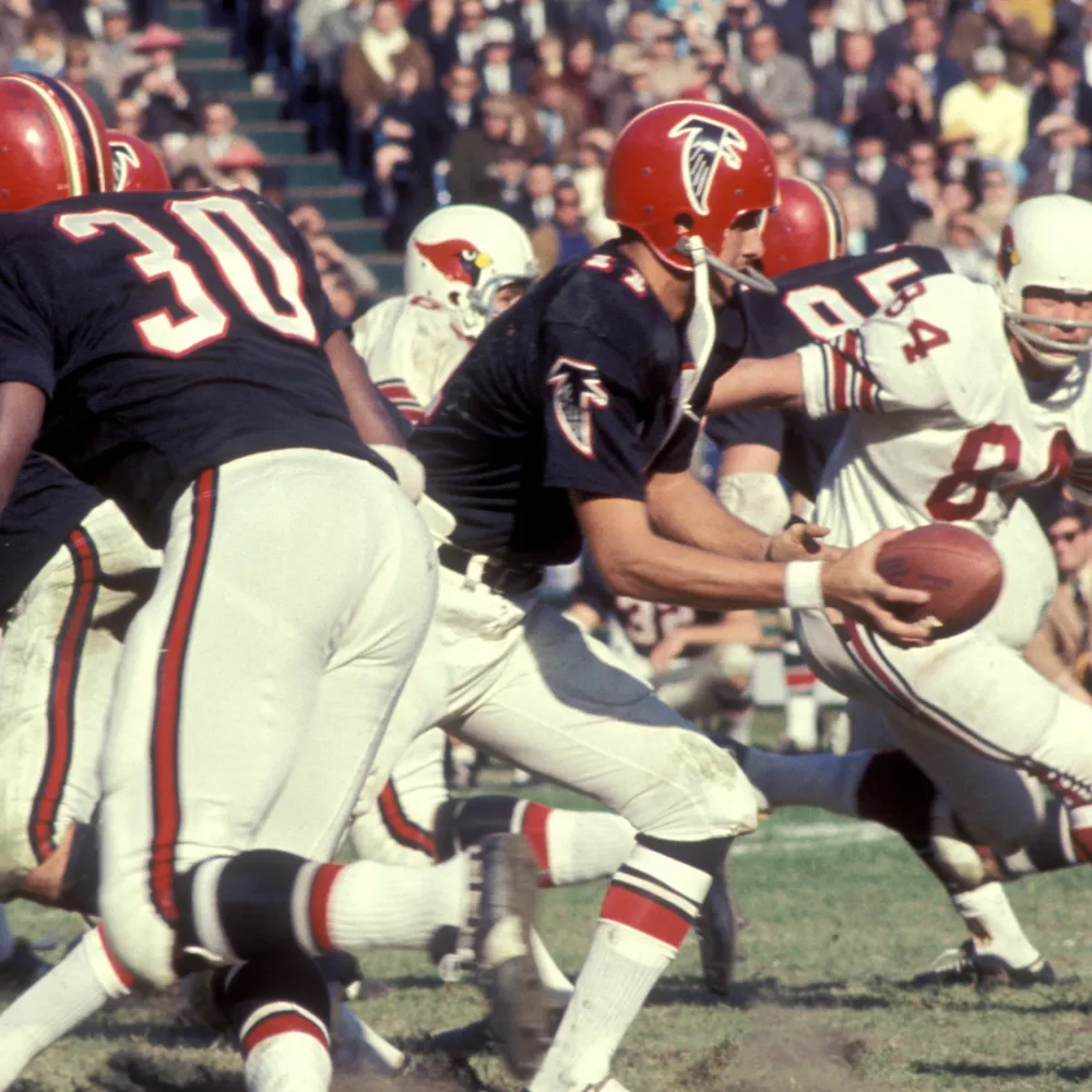 Football game action with Atlanta Falcons player in black jersey and red helmet holding football amid opponents in white Cardinals uniforms.