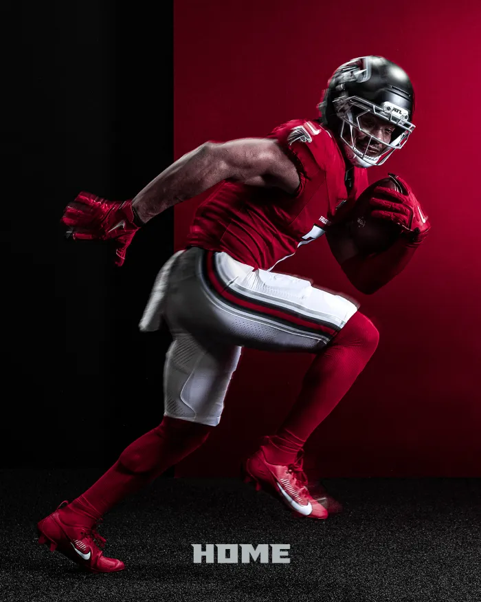 Football player in red jersey, white pants, and black helmet running with football against a black and red background.