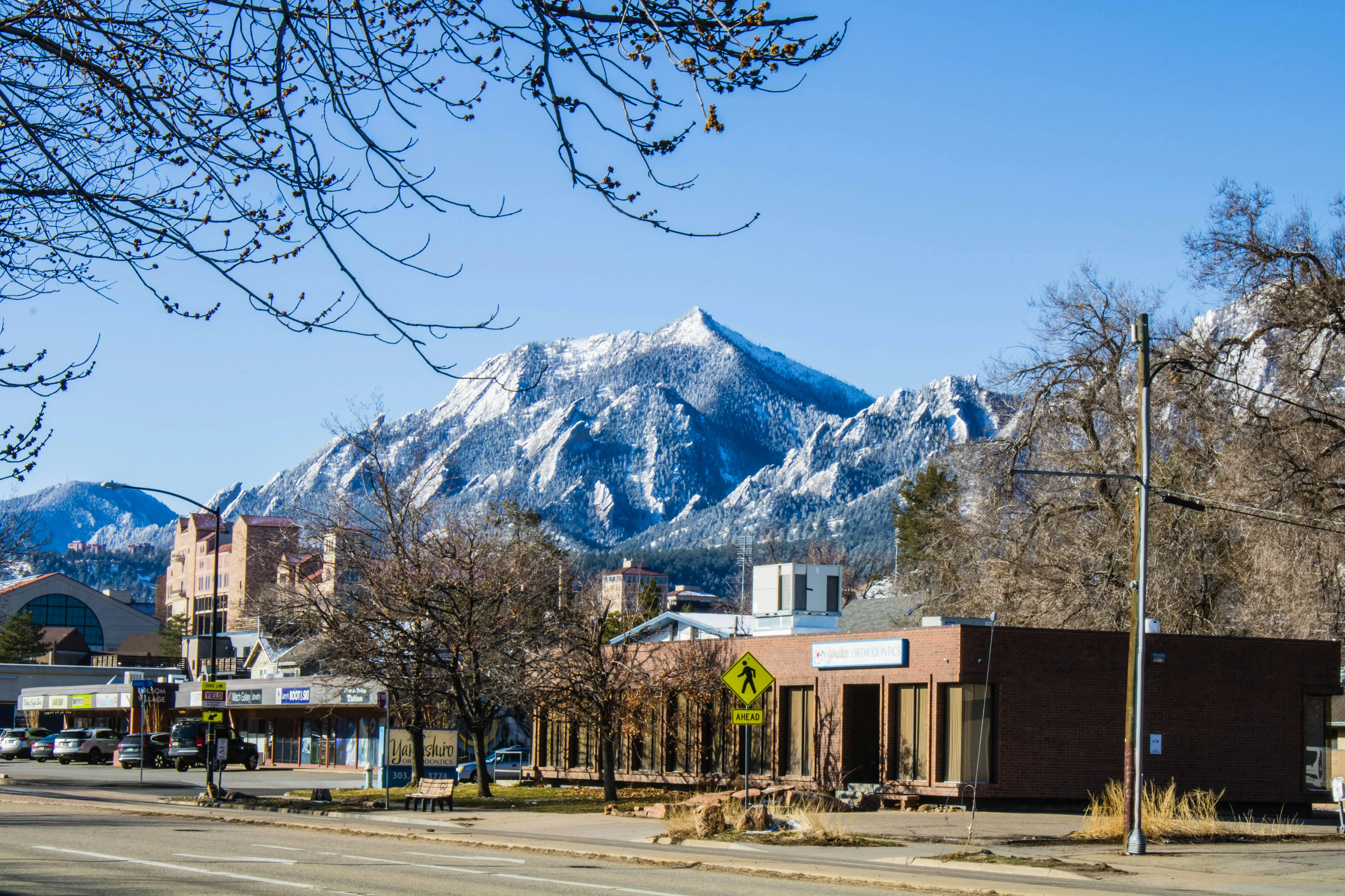 Snow-capped mountain overlooking a small town street with commercial buildings and bare trees under a clear blue sky.