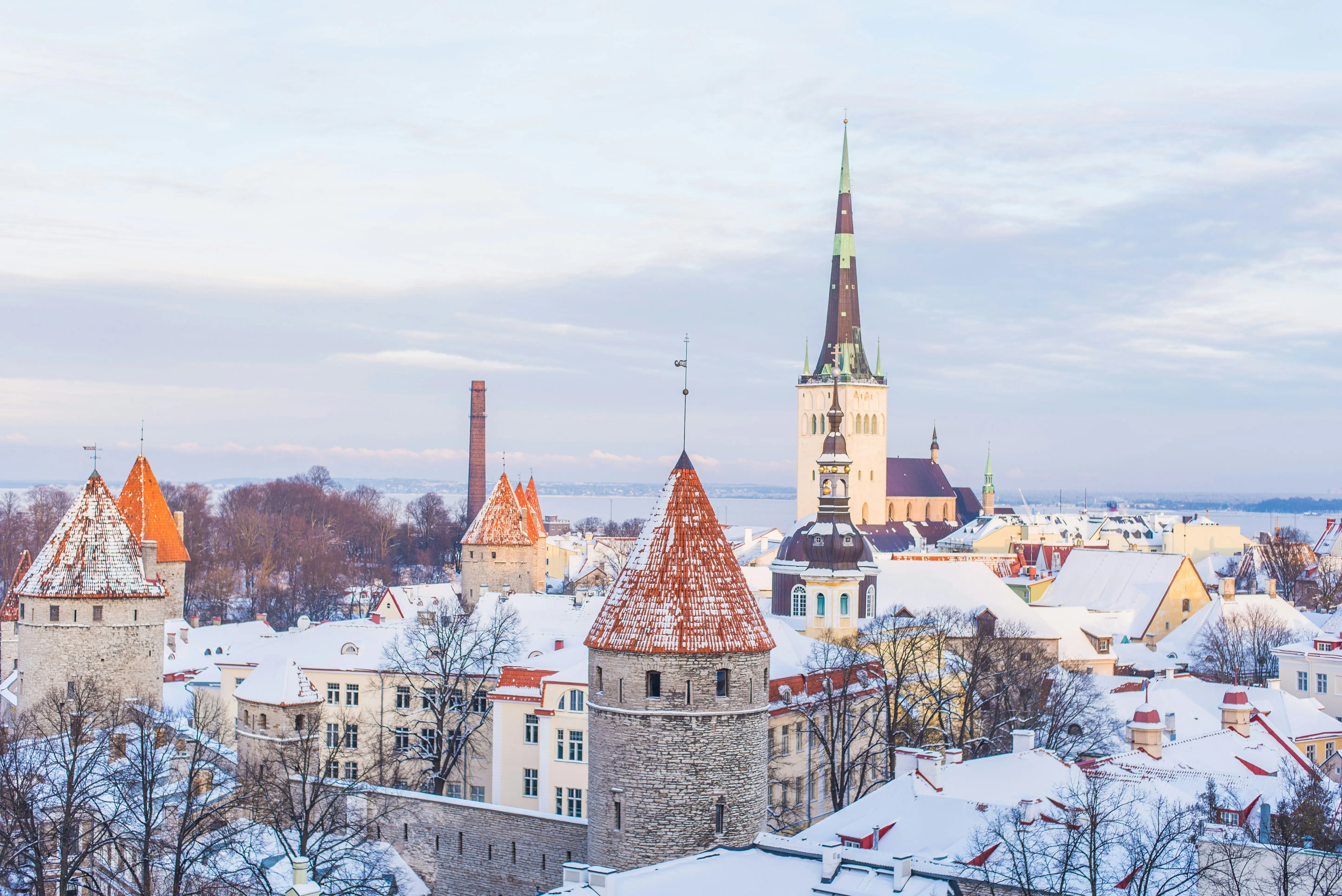 Winter view of Tallinn old town with snow-covered rooftops, medieval stone towers, and a tall church spire under a cloudy sky.