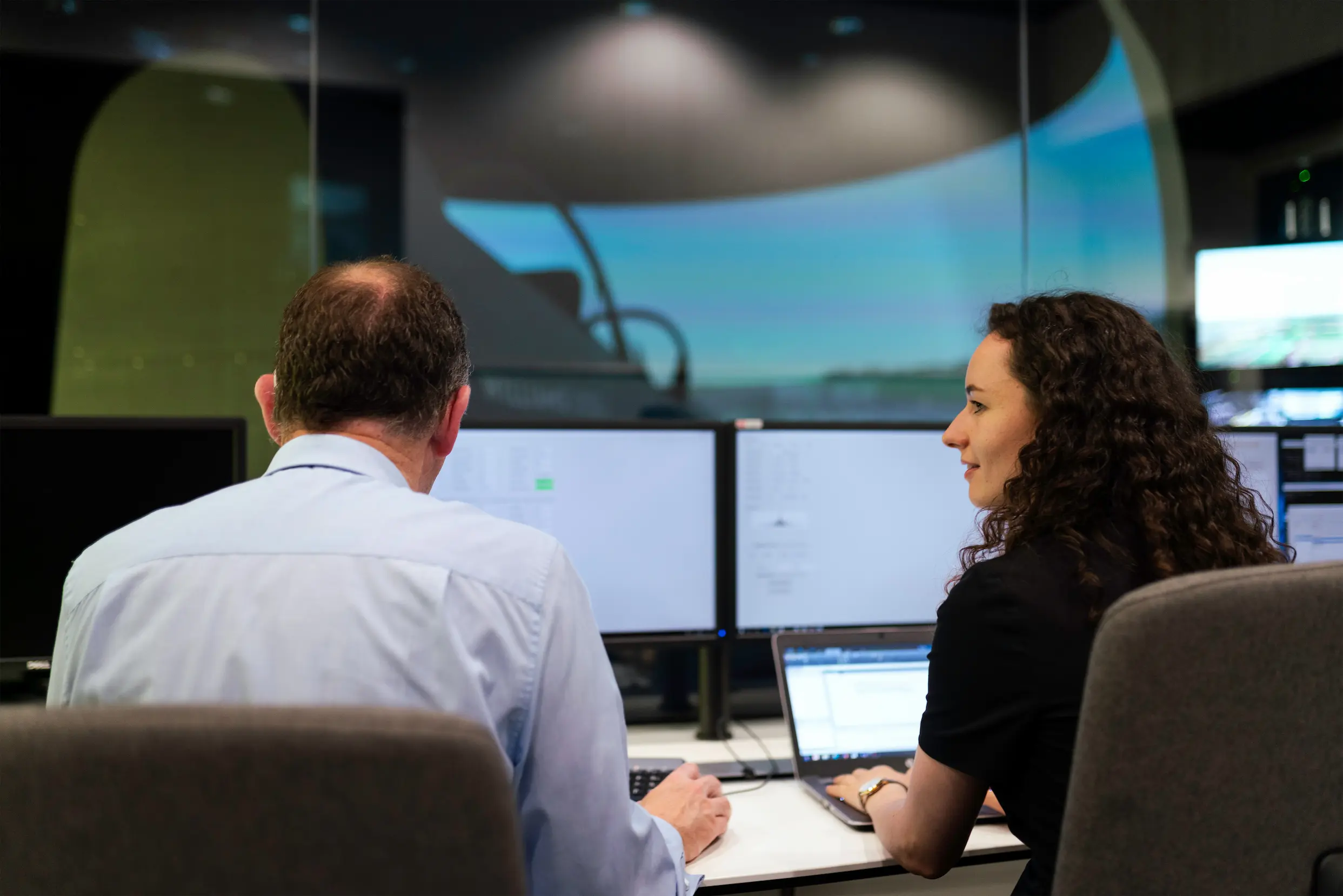 Two professionals working at a desk with multiple computer monitors and a laptop in a modern office setting.