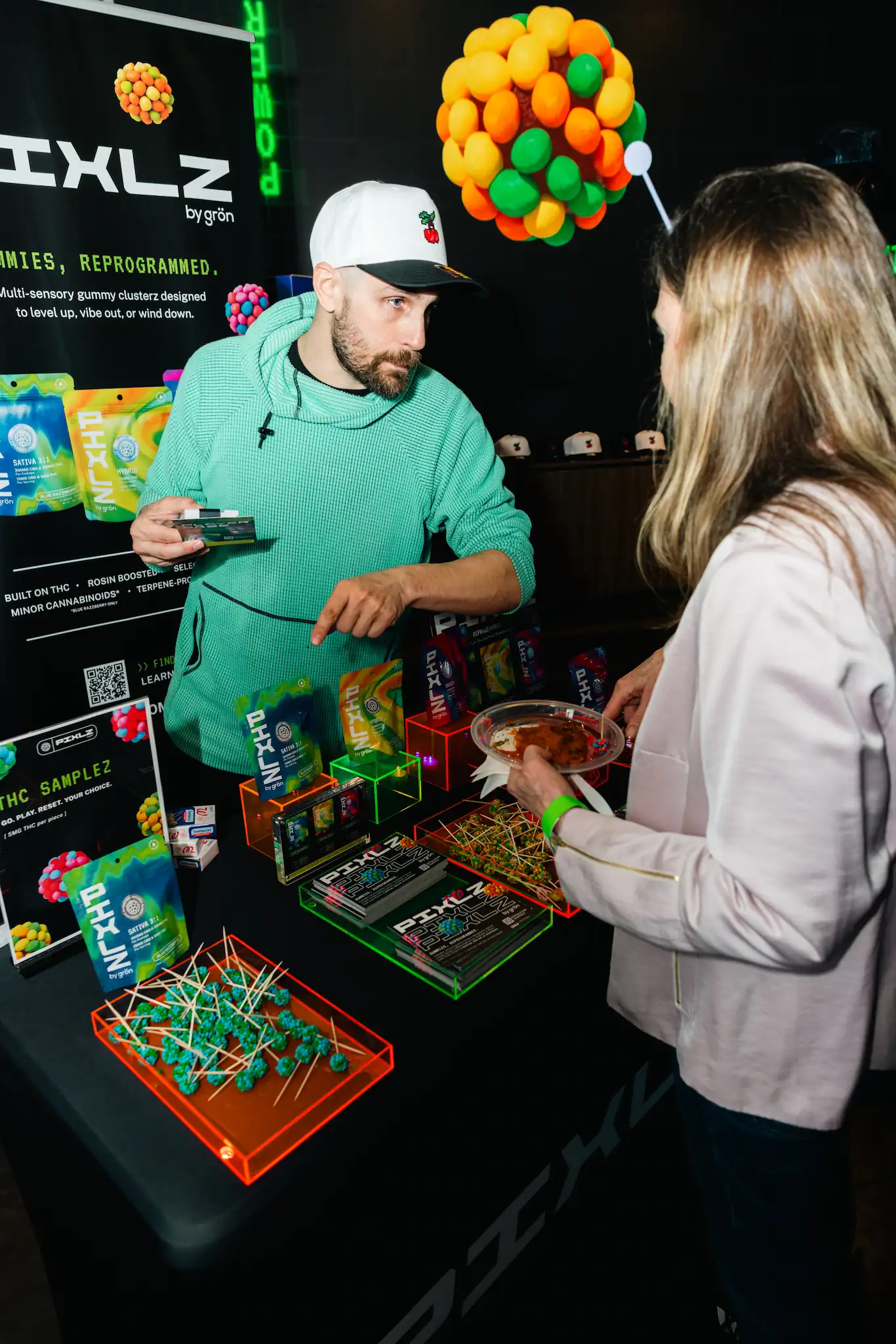 Man in green hoodie explains Pixlz gummy products to a woman at a promotional booth.