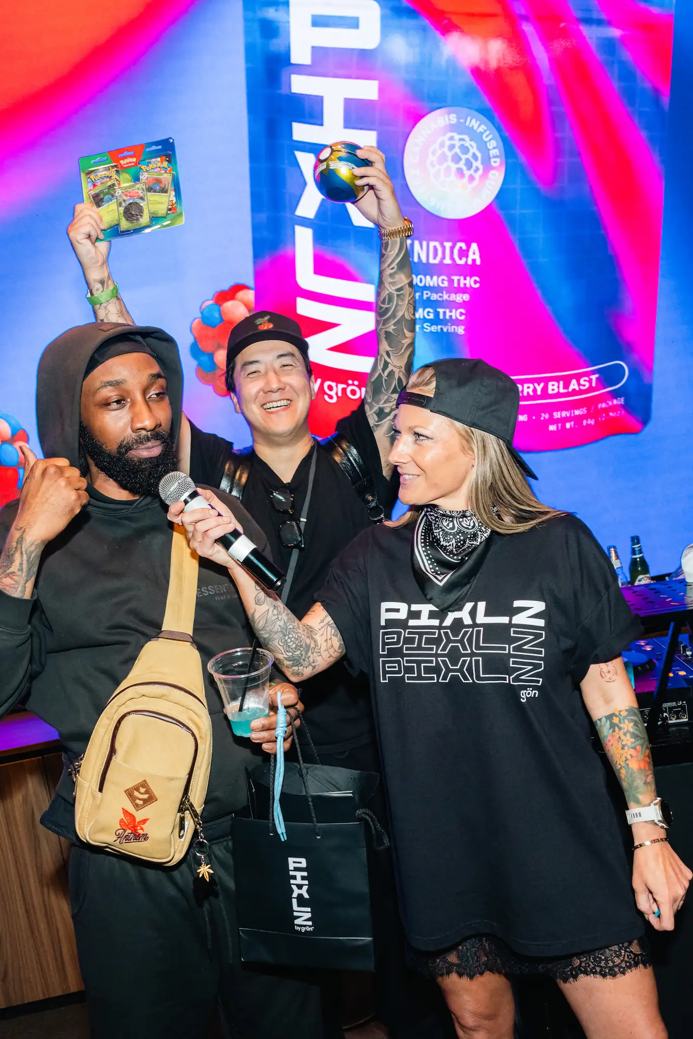 Three people celebrating at a PIXLZ event with microphones, drinks, and product packages in hand.