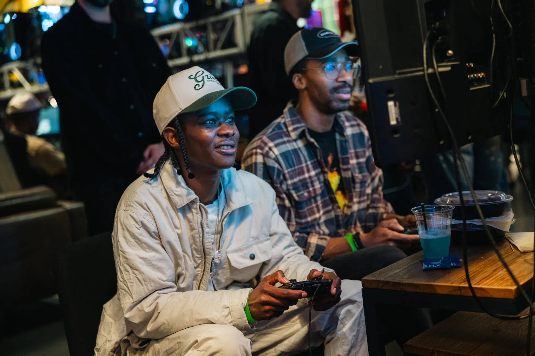 Two men sitting indoors playing video games focused on a TV screen with drinks on a wooden table.