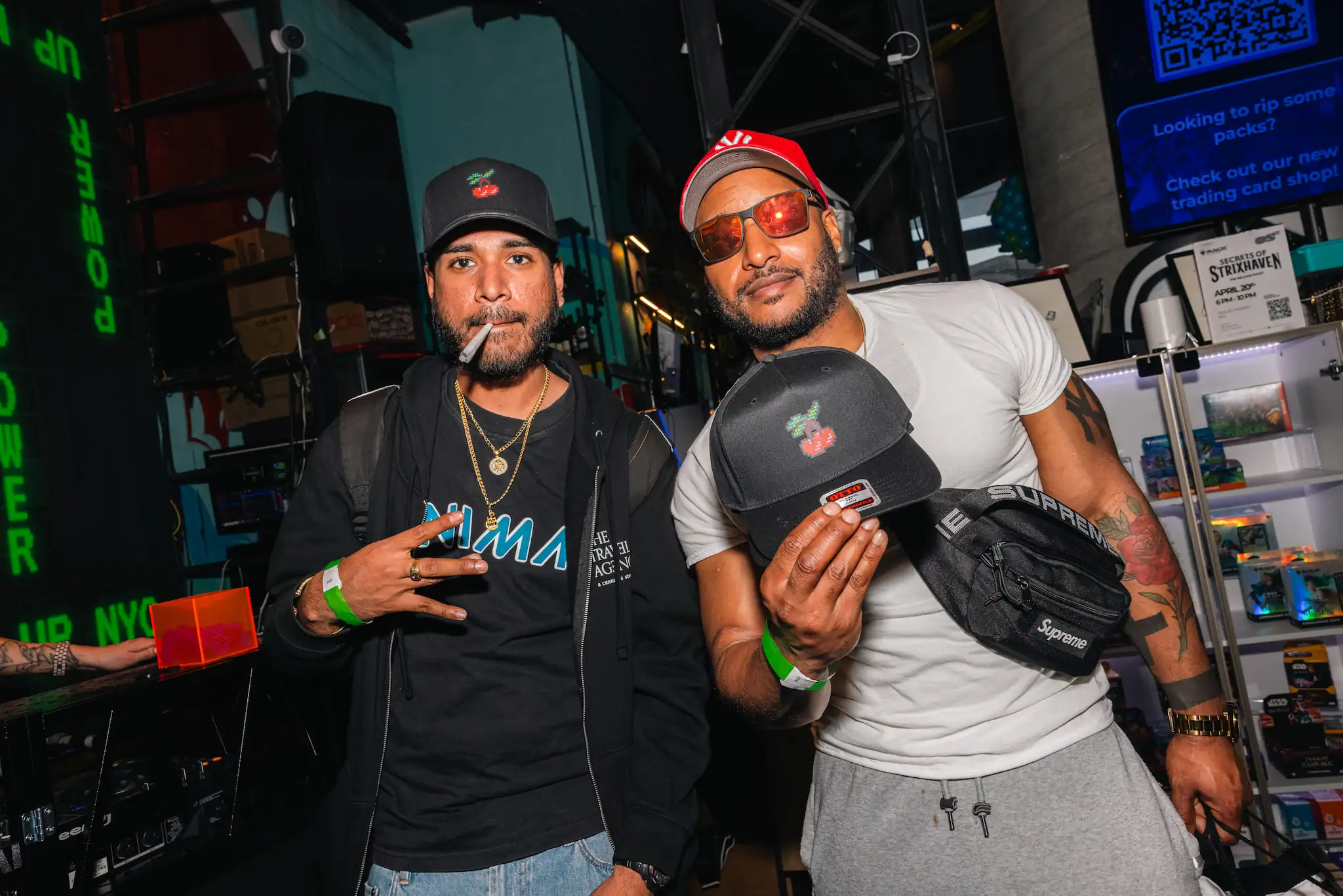 Two men posing at event, one smoking and the other holding a black hat with a cherry logo.