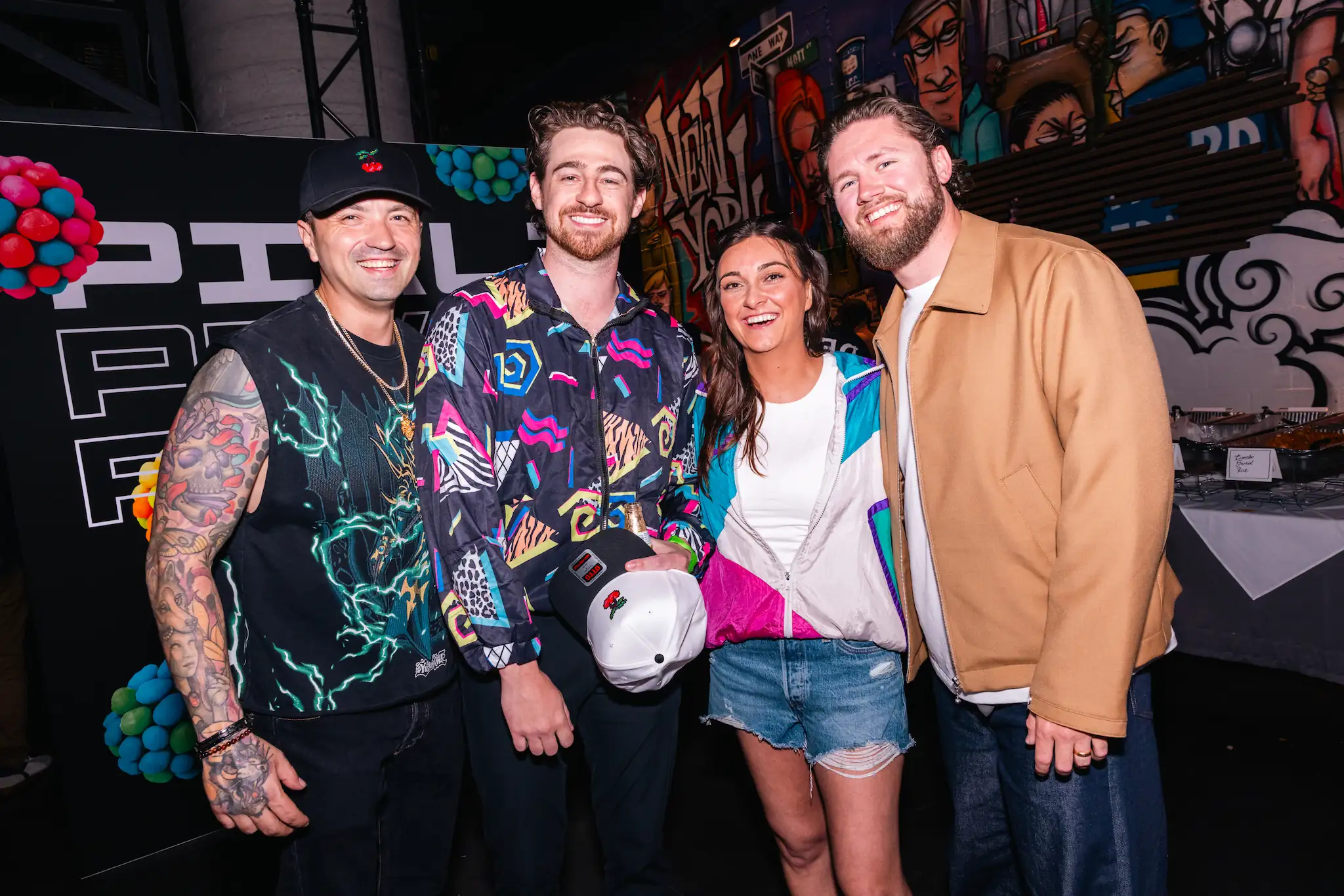 Four smiling people posing together in colorful and casual outfits at an indoor event with graffiti wall art.