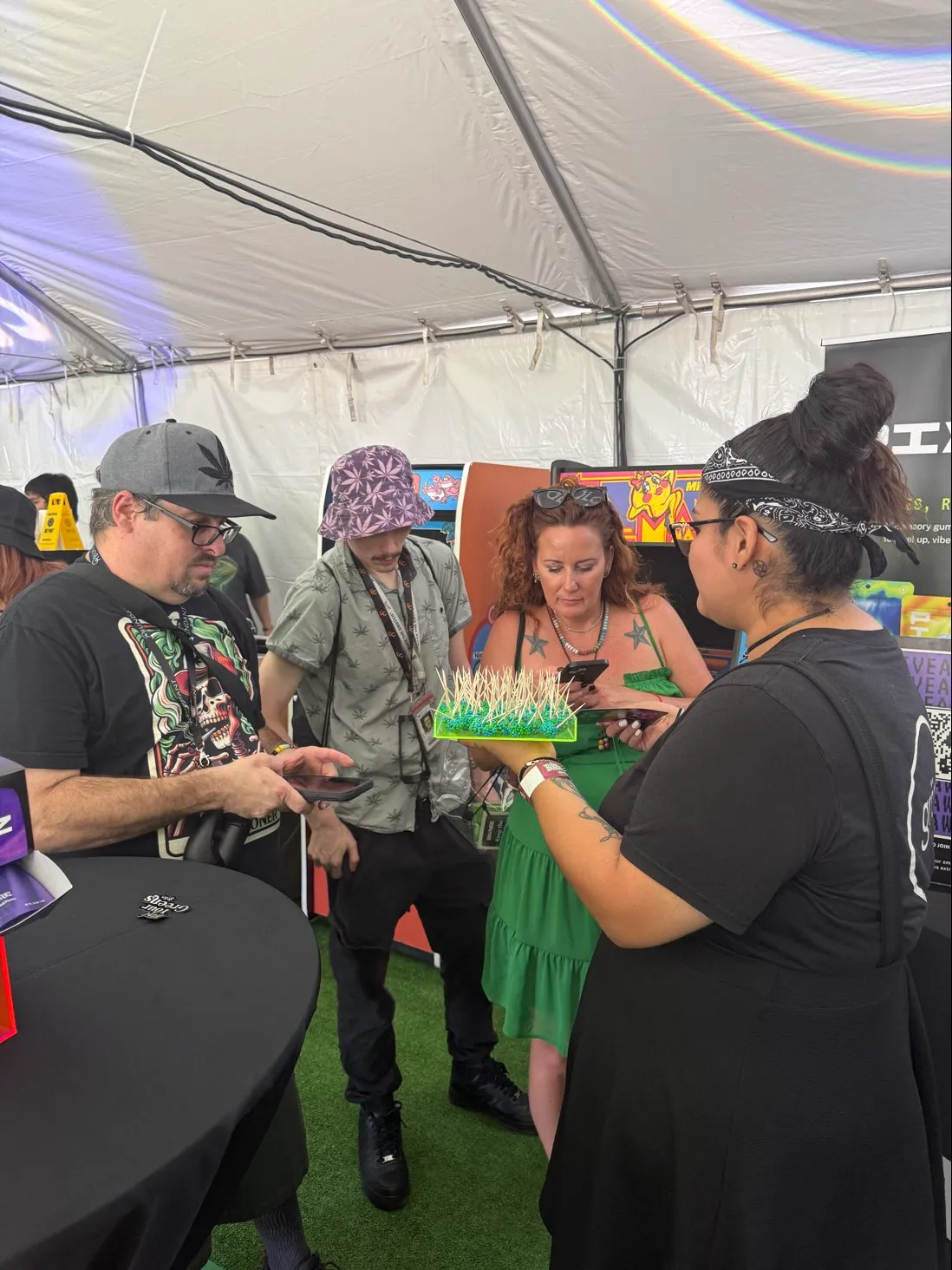Group of four adults inside a tent, one holding a tray of items with toothpicks, others looking at phones.