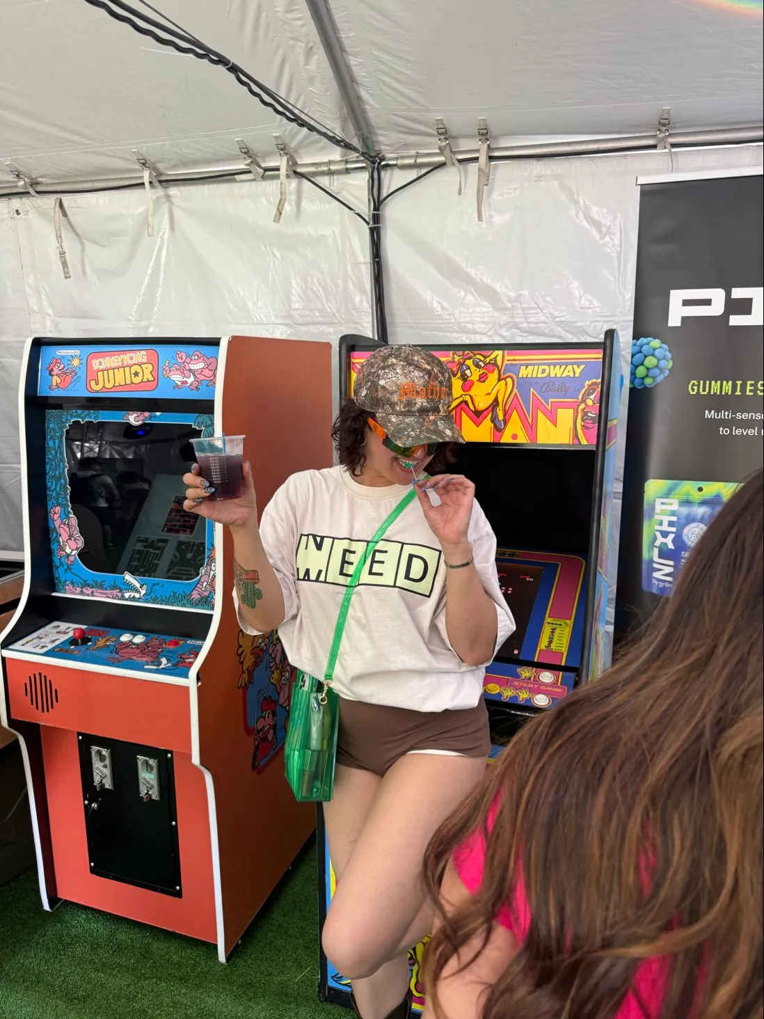 Person in a camouflage hat and WEED shirt drinking with straw in front of arcade machines.