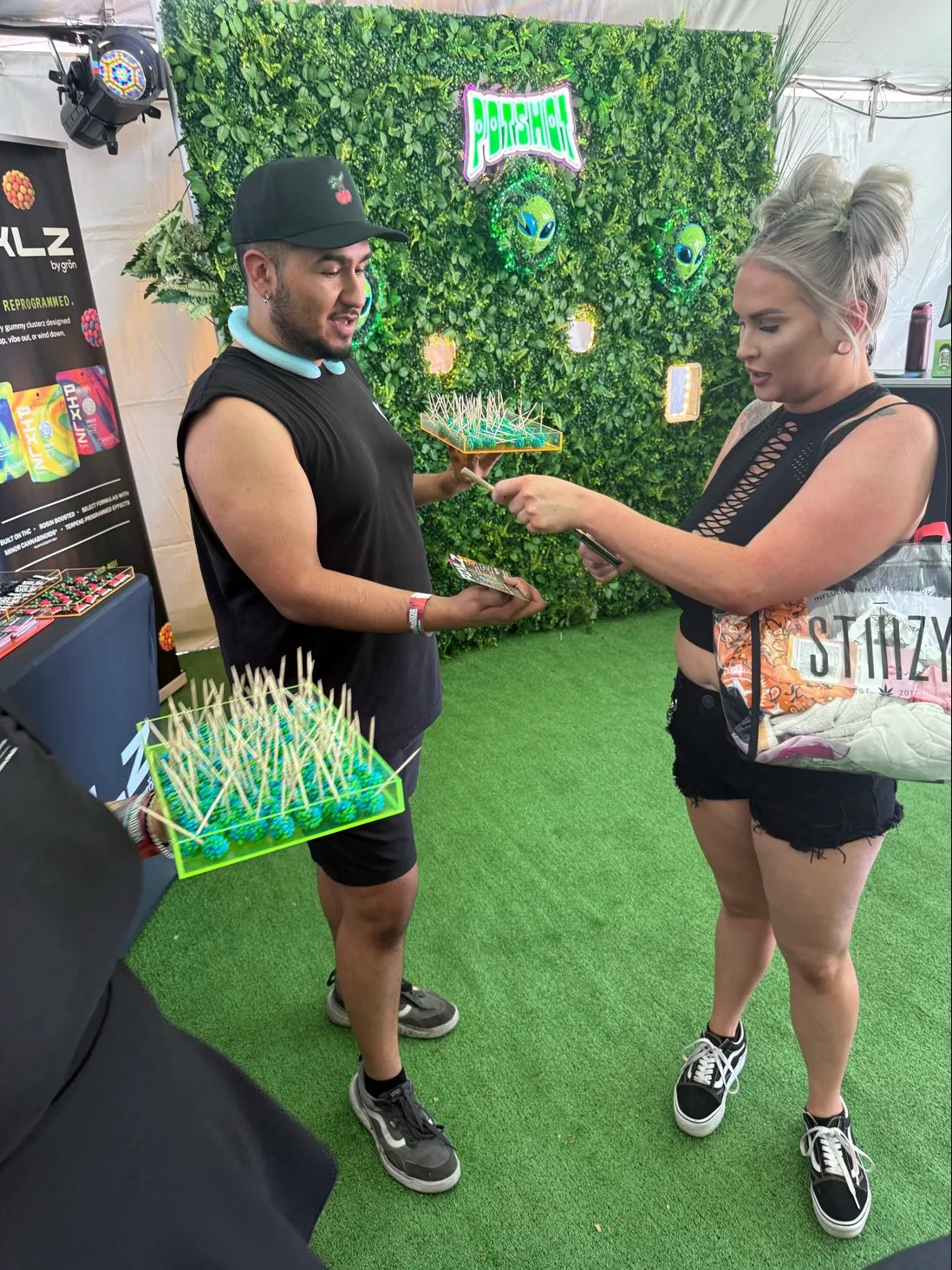 Man holding trays of toothpicked PIXLZ candy offers a woman a sample at an outdoor booth with green wall.