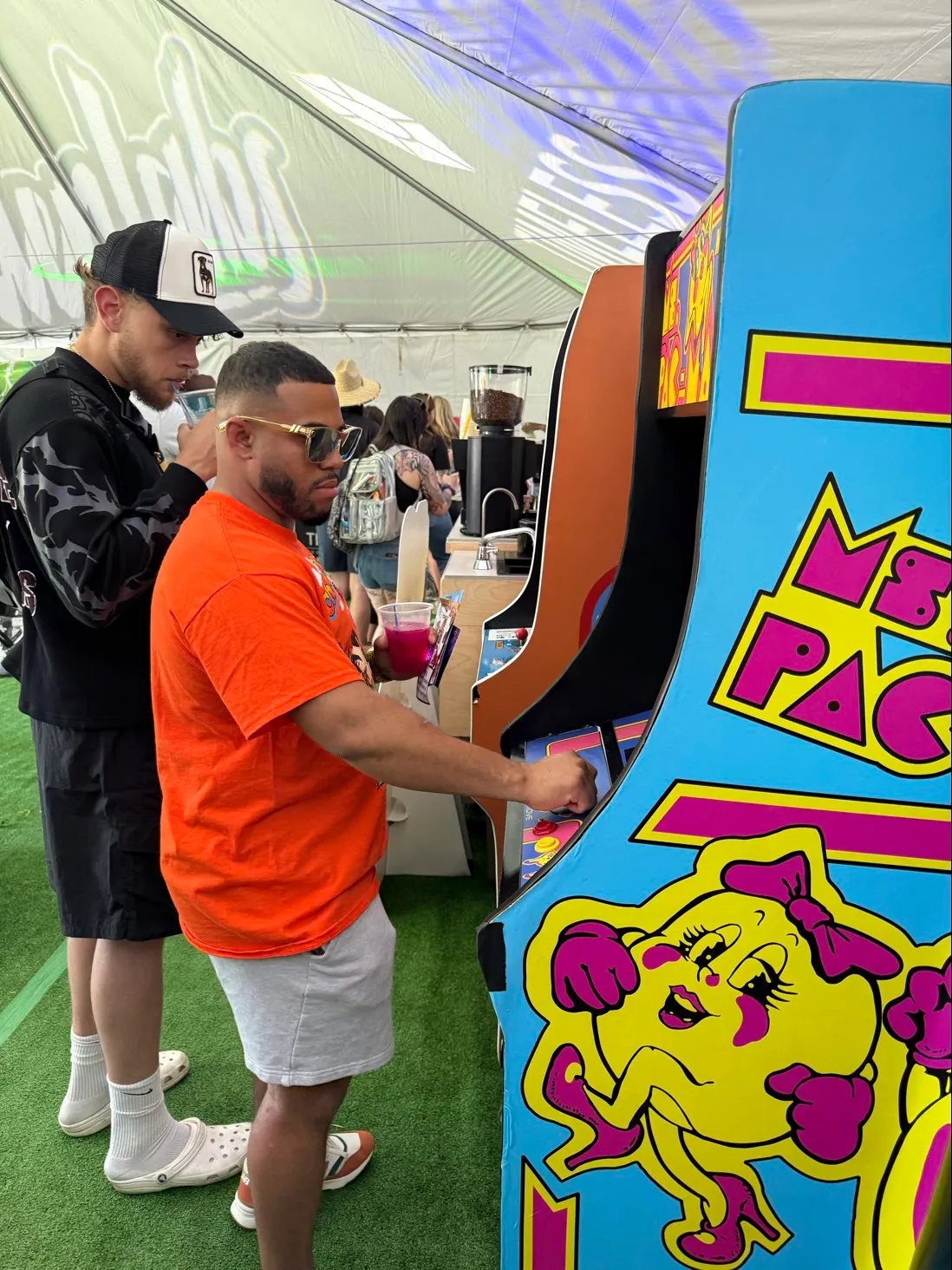 Two men playing vintage arcade games under a tent with one wearing an orange shirt and sunglasses.