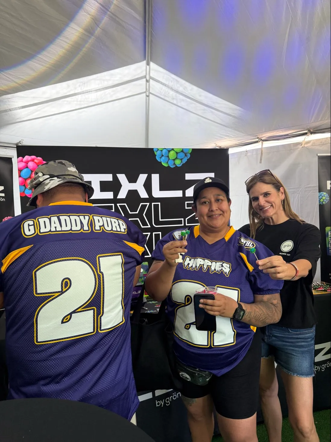 Two people in matching purple jerseys with number 21 and one woman in black shirt smiling under a tent.