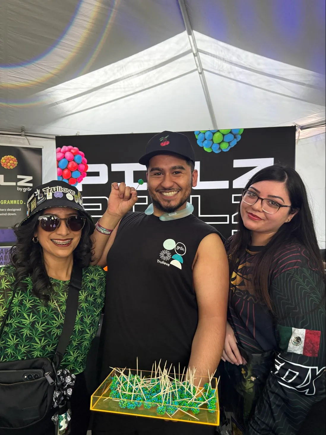 Three smiling people at a booth under a tent, one holding a tray with small green and blue PIXLZ on toothpicks.