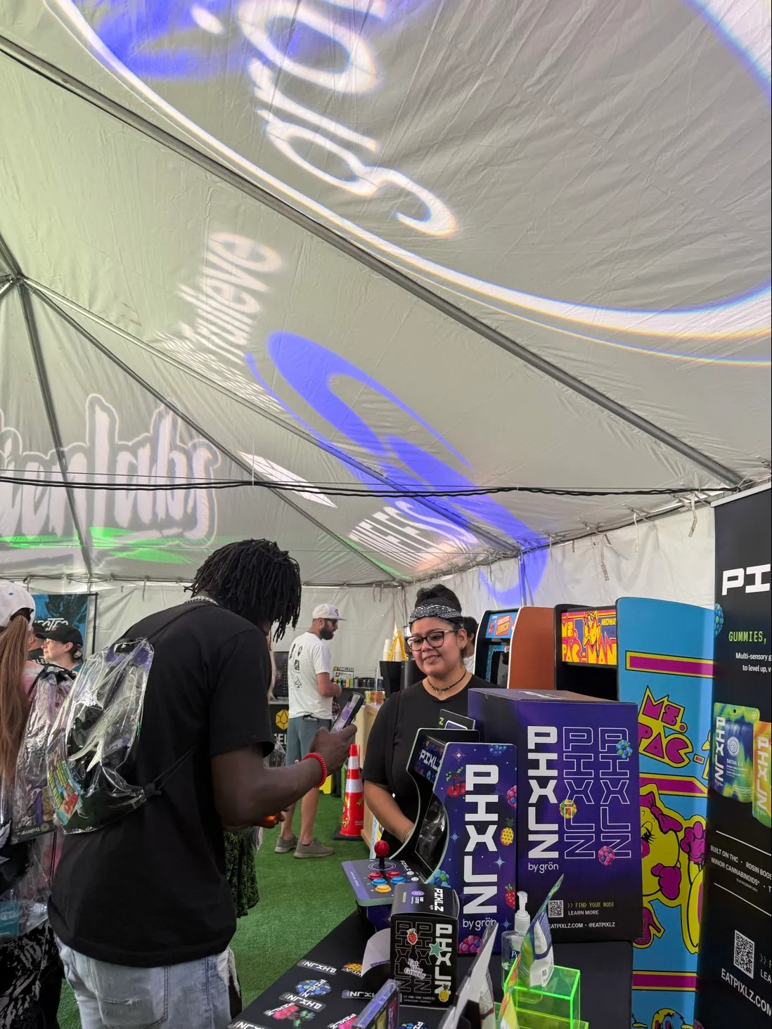 People inside a tent at an event with a retro Ms. Pac-Man arcade game and Pixlz display booth.
