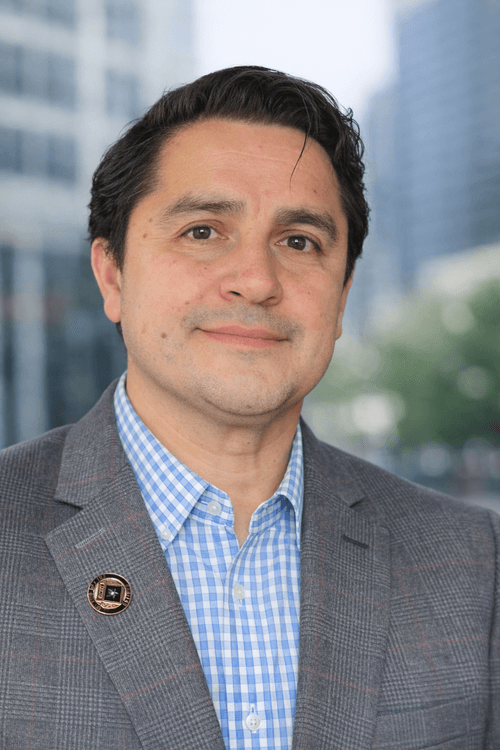 Man in a gray checkered blazer and blue checkered shirt smiling with a blurred cityscape background.