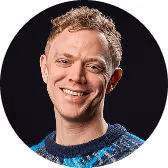 Smiling man with short curly hair wearing a patterned sweater on a black background.
