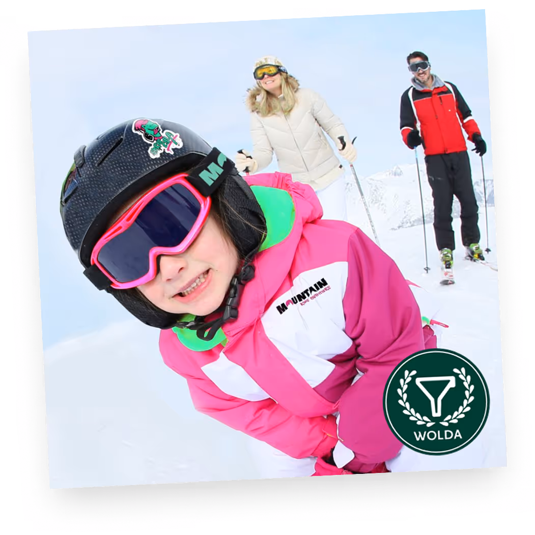 Child in pink ski outfit smiling with two adults skiing in snowy mountain background.