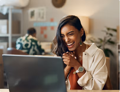 Smiling woman excitedly looks at her laptop screen in a cozy office setting.
