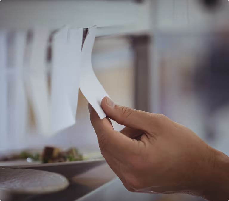 Hand holding a kitchen order ticket hanging on a metal rack above plates.