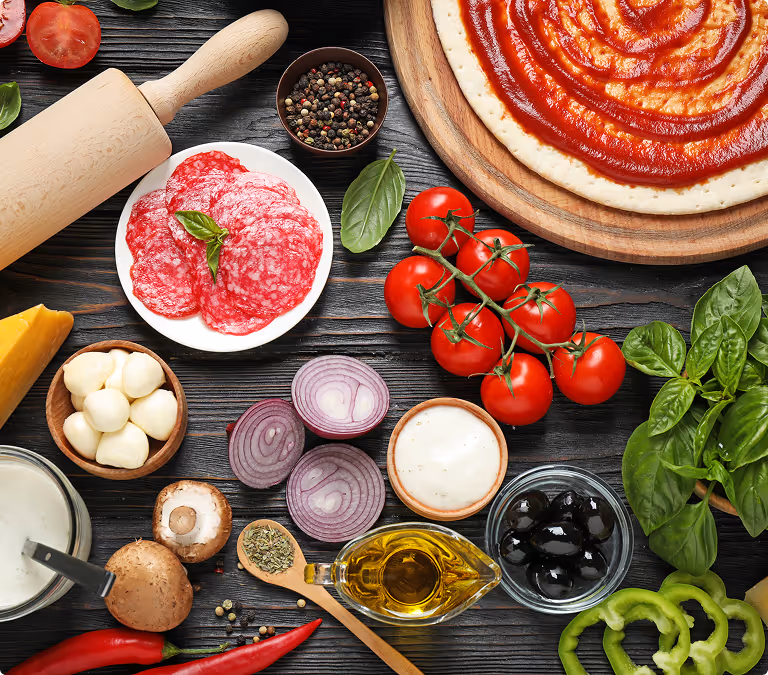 Ingredients for making pizza including dough with sauce, tomatoes, basil, salami, onions, olives, cheese, and olive oil.