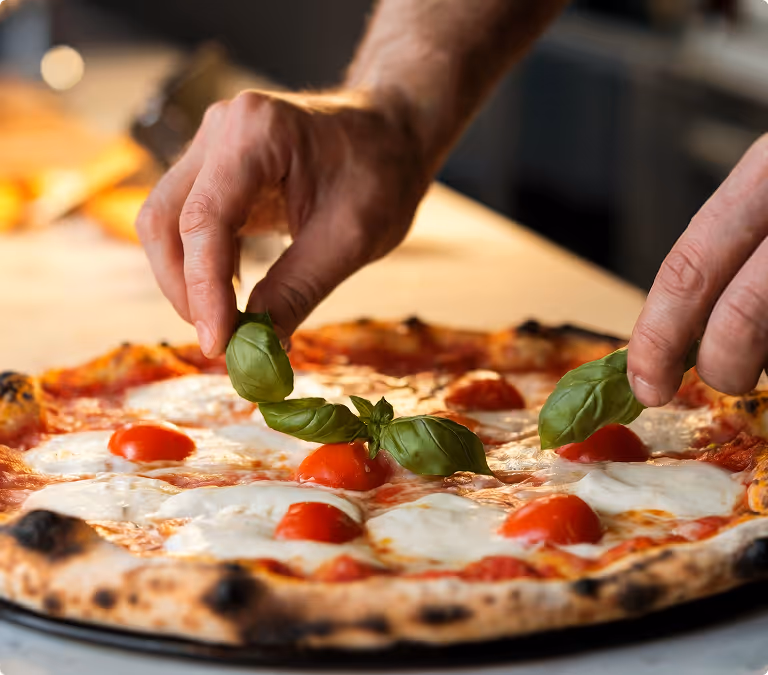 Hands placing fresh basil leaves on a cheese and cherry tomato pizza with a charred crust.