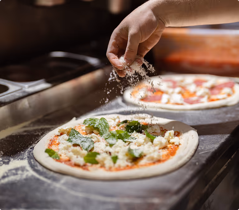 Hand sprinkling cheese over spinach and cheese pizza dough with another pizza in the background.