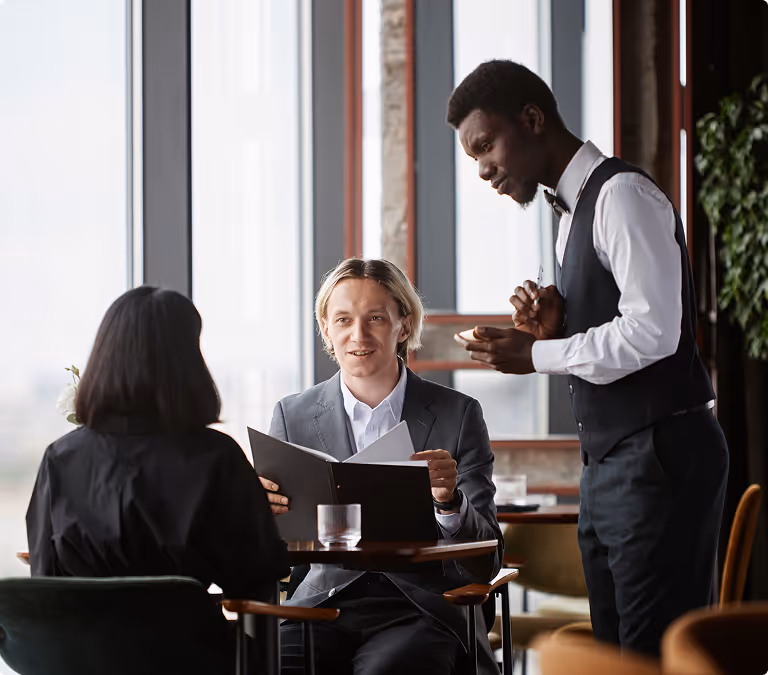 Waiter in vest taking order from a couple seated at a table in a restaurant with large windows.
